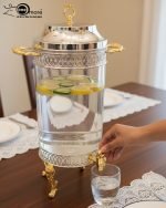 Luxury stainless steel and clear glass beverage dispenser with ornate gold-tone base and handles, available in 7-liter and 10-liter sizes, shown serving infused water at an elegant event.