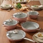 Falez 9-piece pink ceramic granite cookware set featuring a fry pan tossing vegetables, a shallow casserole, and three pots with glass lids, arranged on a kitchen counter and dining table.
