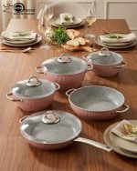 Falez 9-piece pink ceramic granite cookware set featuring a fry pan tossing vegetables, a shallow casserole, and three pots with glass lids, arranged on a kitchen counter and dining table.