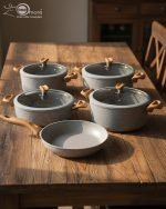 Falez 9-piece grey granite non-stick cookware set displaying a frying pan, shallow casserole, and deep pots with glass lids and wooden-style handles on a kitchen countertop.