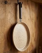 Falez non-stick granite crepe pan in beige speckled finish, shown cooking a golden-brown crepe on a gas stove with a wooden spreader. Option 2: Person flipping a crepe in a Falez granite coated pan, demonstrating non-stick surface and low-oil cooking capabilities.