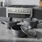 Set of One More non-stick granite casserole pots in 24cm, 26cm, and 28cm sizes with glass lids and ergonomic silicone handles, displayed on a modern kitchen counter.