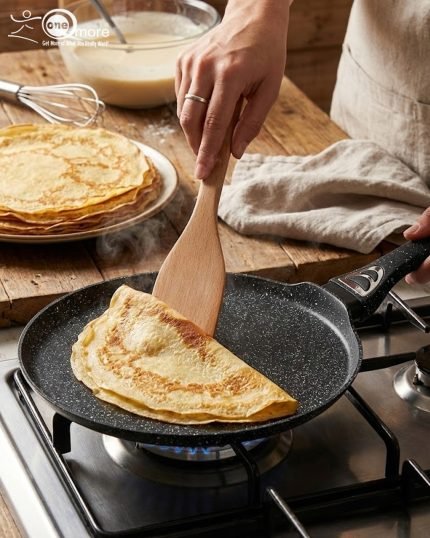 Non-stick 24cm granite crepe pan by One More in black speckled texture, showing a pancake being flipped with a wooden spatula on a gas stove.