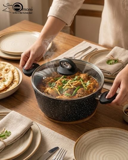 One More 24cm black granite non-stick cooking pot with glass lid and heat-resistant silicone handles, shown preparing healthy low-oil food.