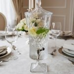 A luxurious 4.5-liter crystal-cut glass beverage dispenser by One More, featuring an intricate diamond pattern and silver spigot, displayed on a formal dining table with lemon-infused water.