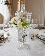 A luxurious 4.5-liter crystal-cut glass beverage dispenser by One More, featuring an intricate diamond pattern and silver spigot, displayed on a formal dining table with lemon-infused water.