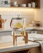 A One More brand 3.6 liter ribbed glass water dispenser filled with lemon slices and ice, featuring a gold tap, a brown leather handle, and a heart-shaped lid knob, resting on a light wood bamboo stand in a modern kitchen and living room setting.