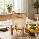 Elegant 5.5L ribbed glass beverage dispenser featuring a gold tap, leather handle, and heart-shaped gold lid knob, displayed on a wooden bamboo stand in a bright, modern dining setting.
