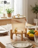 Elegant 5.5L ribbed glass beverage dispenser featuring a gold tap, leather handle, and heart-shaped gold lid knob, displayed on a wooden bamboo stand in a bright, modern dining setting.