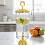 Elegant 3.5L clear glass water dispenser with luxury gold base, gold lid with ring handle, and a gold spigot, filled with infused water and citrus slices on a marble kitchen countertop.