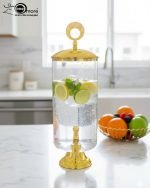 Elegant 3.5L clear glass water dispenser with luxury gold base, gold lid with ring handle, and a gold spigot, filled with infused water and citrus slices on a marble kitchen countertop.