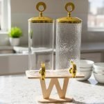 A luxury dual glass beverage dispenser set by One More featuring a 3.3L smooth jar and a 3.3L hammered glass jar, equipped with gold metallic spigots and lids, displayed on a stylish W-shaped wooden stand on a decorated dinner table.