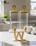 A luxury dual glass beverage dispenser set by One More featuring a 3.3L smooth jar and a 3.3L hammered glass jar, equipped with gold metallic spigots and lids, displayed on a stylish W-shaped wooden stand on a decorated dinner table.