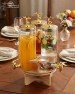 Elegant triple glass beverage dispenser set on a rotating wooden stand, featuring gold-toned taps and decorative deer figurines on lids, shown in various settings including a dinner party and outdoor event.