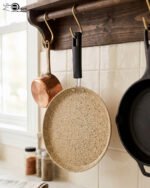 Falez non-stick granite crepe pan in beige speckled finish, shown cooking a golden-brown crepe on a gas stove with a wooden spreader. Option 2: Person flipping a crepe in a Falez granite coated pan, demonstrating non-stick surface and low-oil cooking capabilities.