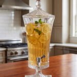Elegant One More 4.5-liter ribbed glass beverage dispenser filled with orange juice and ice, featuring a silver tap and matching glass lid on a wooden table.