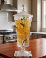 Elegant One More 4.5-liter ribbed glass beverage dispenser filled with orange juice and ice, featuring a silver tap and matching glass lid on a wooden table.