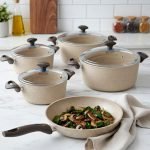 Falez 9-piece beige granite non-stick cookware set arranged on a kitchen counter, featuring a fry pan, shallow casserole, and three deep pots with glass lids, showcasing healthy low-oil cooking.