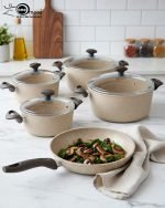 Falez 9-piece beige granite non-stick cookware set arranged on a kitchen counter, featuring a fry pan, shallow casserole, and three deep pots with glass lids, showcasing healthy low-oil cooking.