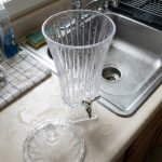 Elegant One More 4.5-liter ribbed glass beverage dispenser filled with orange juice and ice, featuring a silver tap and matching glass lid on a wooden table.