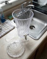 Elegant One More 4.5-liter ribbed glass beverage dispenser filled with orange juice and ice, featuring a silver tap and matching glass lid on a wooden table.