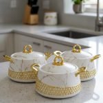 Three One More branded insulated hotpots in black and white marble finishes with gold honeycomb detailing and gold handles, shown in a kitchen and dining room setting with steaming food.