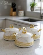 Three One More branded insulated hotpots in black and white marble finishes with gold honeycomb detailing and gold handles, shown in a kitchen and dining room setting with steaming food.