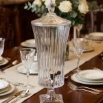 An elegant 4.5-liter ribbed glass beverage dispenser filled with pink lemonade and lime slices, standing on a wooden dining table with crystal glasses and floral arrangements in a cinematic lit background.