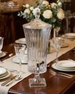 An elegant 4.5-liter ribbed glass beverage dispenser filled with pink lemonade and lime slices, standing on a wooden dining table with crystal glasses and floral arrangements in a cinematic lit background.