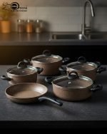 Falez 9-piece brown speckled granite non-stick cookware set displayed on a kitchen counter, featuring a fry pan, shallow casserole, and three deep pots with glass lids - Made in Turkey.