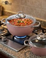 Falez 9-piece pink ceramic granite cookware set featuring a fry pan tossing vegetables, a shallow casserole, and three pots with glass lids, arranged on a kitchen counter and dining table.
