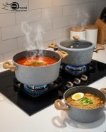 Falez 9-piece grey granite non-stick cookware set displaying a frying pan, shallow casserole, and deep pots with glass lids and wooden-style handles on a kitchen countertop.