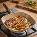 Falez square granite non-stick grill pan cooking grilled chicken breast and vegetables with perfect char marks, featuring a beige speckled finish and ergonomic brown handle.