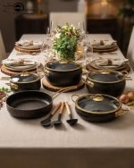 One More 12-piece black granite non-stick cookware set displayed on a wooden table, featuring gold handles, glass lids, a fry pan, shallow casserole, deep pots, and cooking spoons.