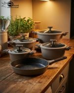 UCC LIFE 9-piece non-stick granite cookware set arranged on a wooden kitchen counter, featuring a frying pan, shallow casserole, and three pots with glass lids in a speckled grey finish.