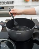 Set of One More non-stick granite casserole pots in 24cm, 26cm, and 28cm sizes with glass lids and ergonomic silicone handles, displayed on a modern kitchen counter.