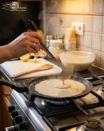 Non-stick 24cm granite crepe pan by One More in black speckled texture, showing a pancake being flipped with a wooden spatula on a gas stove.