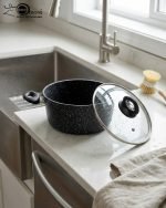 One More 24cm black granite non-stick cooking pot with glass lid and heat-resistant silicone handles, shown preparing healthy low-oil food.