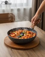 One More grey granite non-stick wok pan with a single long wooden-style handle, cooking shrimp and vegetables on a stove, displaying stone texture.