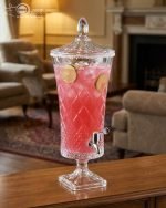 A luxurious 4.5-liter crystal-cut glass beverage dispenser by One More, featuring an intricate diamond pattern and silver spigot, displayed on a formal dining table with lemon-infused water.