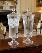 An elegant 4.5-liter ribbed glass beverage dispenser filled with pink lemonade and lime slices, standing on a wooden dining table with crystal glasses and floral arrangements in a cinematic lit background.