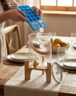 A One More brand 3.6 liter ribbed glass water dispenser filled with lemon slices and ice, featuring a gold tap, a brown leather handle, and a heart-shaped lid knob, resting on a light wood bamboo stand in a modern kitchen and living room setting.