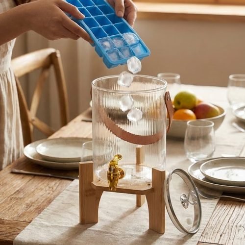 A One More brand 3.6 liter ribbed glass water dispenser filled with lemon slices and ice, featuring a gold tap, a brown leather handle, and a heart-shaped lid knob, resting on a light wood bamboo stand in a modern kitchen and living room setting.