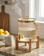 Elegant 5.5L ribbed glass beverage dispenser featuring a gold tap, leather handle, and heart-shaped gold lid knob, displayed on a wooden bamboo stand in a bright, modern dining setting.