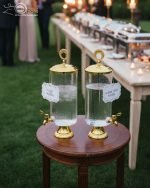 Elegant 3.5L clear glass water dispenser with luxury gold base, gold lid with ring handle, and a gold spigot, filled with infused water and citrus slices on a marble kitchen countertop.