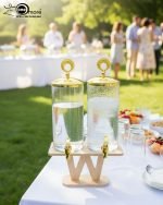 A luxury dual glass beverage dispenser set by One More featuring a 3.3L smooth jar and a 3.3L hammered glass jar, equipped with gold metallic spigots and lids, displayed on a stylish W-shaped wooden stand on a decorated dinner table.