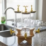 Elegant triple glass beverage dispenser set on a rotating wooden stand, featuring gold-toned taps and decorative deer figurines on lids, shown in various settings including a dinner party and outdoor event.