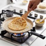 Falez non-stick granite crepe pan in beige speckled finish, shown cooking a golden-brown crepe on a gas stove with a wooden spreader. Option 2: Person flipping a crepe in a Falez granite coated pan, demonstrating non-stick surface and low-oil cooking capabilities.