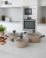 Falez 9-piece beige granite non-stick cookware set arranged on a kitchen counter, featuring a fry pan, shallow casserole, and three deep pots with glass lids, showcasing healthy low-oil cooking.