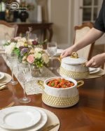 Three One More branded insulated hotpots in black and white marble finishes with gold honeycomb detailing and gold handles, shown in a kitchen and dining room setting with steaming food.